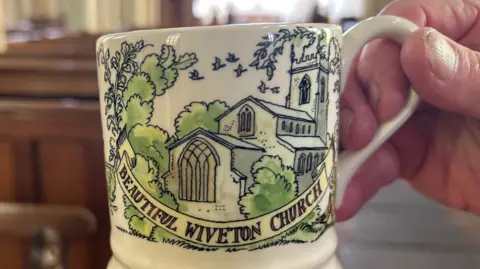 Immy Lee/BBC A painting of the church surrounded by trees and with the words: Beautiful Wiveton Church, is held up in the church. 