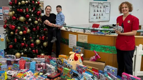 Polly Hurst Polly, a woman, wearing a black shirt and black trousers, holding her son in her arms, standing in front of a Christmas tree with boxes of presents in front of them and a member of the children's ward, a lady in red shirt smiling for camera with them.