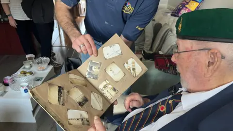 George Carden/BBC Joseph Cheshire wearing his green beret and smart suit with a red poppy badge sat looking at some of his old pictures in his living room