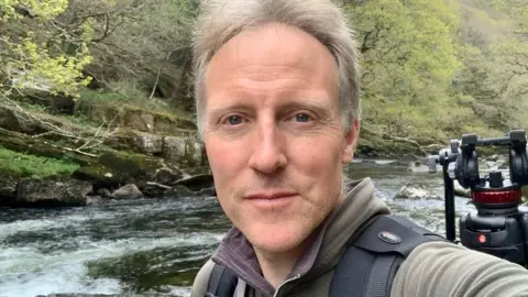 Handout Tim Martin looks into the camera. He does not smile. Behind him a river can be seen with a tripod on his back to the right. He has short, grey hair and is wearing a light green jumper. Black backstraps for a backpack can be seen.