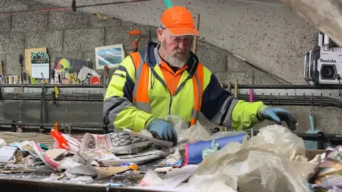 BBC A worker at Mayside Recycling sorts through paper waste. He has a beard, is wearing high-vis and a bright orange hat.