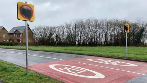 Two 20mph roads signs, one on either side of a road, which have been sprayed painted over covering the number 20.
The signs are placed in a residential area with house and trees surrounding the road