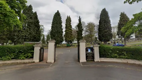 The front gates of a cemetery and crematorium. There are two beige posts with an open black gate. Behind are trees and grass with headstones on it.