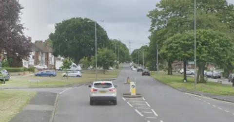 A road with grassy areas and trees on either side. A car is driving away from the camera and other cars are parked on the side of the road.