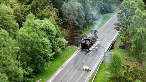 A smouldering burned-out lorry on one side of an empty road. Two firefighters are on the other side of the road
