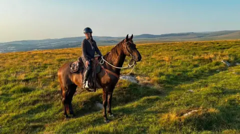 Adventure B4 Dementia A woman on a brown horse. They are on a hill with vast rolling countryside in the background. 
