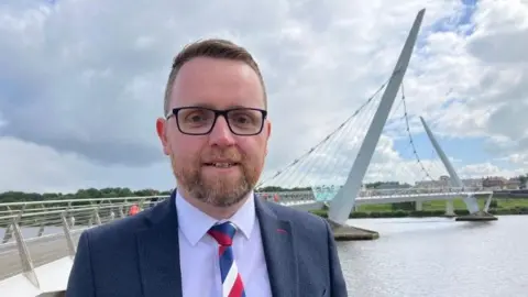 Shows a man with a beard and black-rimmed glasses, a blue jacket, white shirt, red-white-and-blue tie standing in front of the Foyle Bridge.
