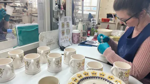 A woman wearing a pink jumper, blue apron, blue gloves and glasses is hand-making a teacup. Several tea cups and bowls are placed on the table next to her. 