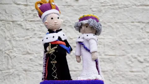 PA Media A woollen post box topper with figures of King Charles and Queen Camilla on top of a red post box