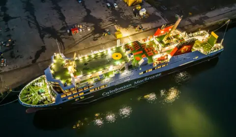 Stephen Lipton An aerial shot of Glen Sannox - a large black and white ship with green decks, lit up at night, moored at a quay beside a large empty industrial plot