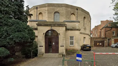 Google A screenshot from Google Street View showing the exterior entrance to Gloucester Crown Court. It is an octagon-shaped sand-coloured building with an arched wooden doorway, bordered by a large tree and a car park on the right.
