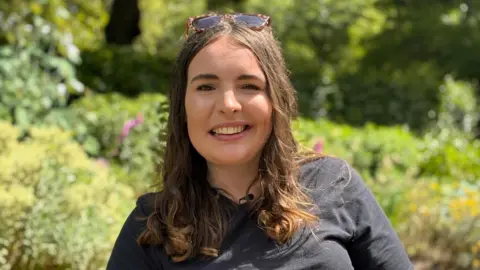 BBC Wales A woman in her late twenties with long brown hair, wearing a black top, smiling at the camera. She's sat in a public park with greenery behind her.