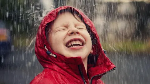 A smiling boy wearing a red raincoat with the hood up, as heavy rain splashes on his face