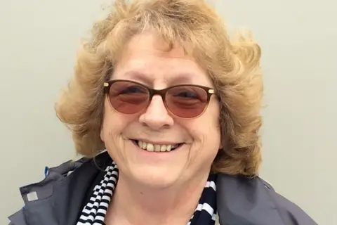 Rosie has ginger hair and wears tinted sunglasses, a grey jacket and a black and white scarf. She is smiling.