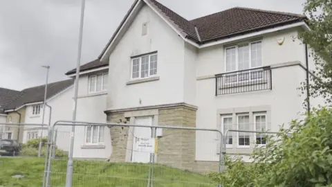 Detached two-storey white house with a tiled roof behind temporary metal safety fencing, viewed from across a grassy front garden on an overcast day. Stone cladding surrounds the ground-floor entrance area, with neighbouring homes, streetlights and trees visible in the background.