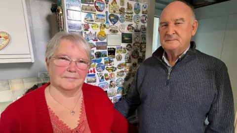 June and Michael Groom, June is wearing a red cardigan and top. She has white hair and clear glasses on. Michael is wearing a blue quarter zip top and has receding white hair. They're standing in their kitchen and behind them is a fridge full of holiday magnets.