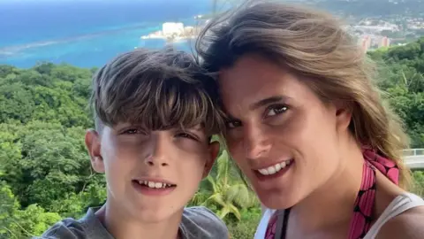 Ellen Roome and her son Jools smile at the camera, they are on holiday and stand at a viewpoint overlooking a forest with a town and beach in the background. It is a bright sunny day.