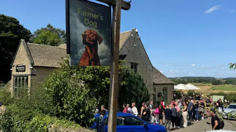 BBC An historic stone pub building under blue skies with rolling fields in the distance. A pub sign with a picture of a fox red labrador on it reads 'The Farmer's Dog'. There is a queue of people outside the pub door. 