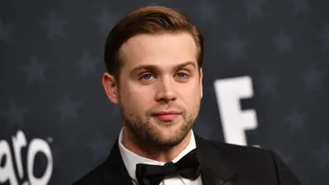 The headshot of Leo Woodall shows him wearing a white shirt with a black suit jacket and bowtie. He stands against a grey background with stars and has short brown hair and green-blue eyes. 