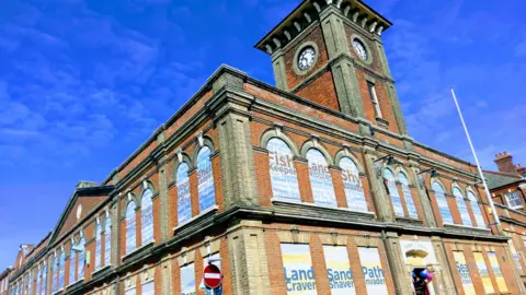 Lowestoft Town Council Lowestoft Town Hall from the outside. It is a large building with a clock tower. All the windows have been covered with identical posters. On the ground floor these read 'Land Craver' and on the top they read 'Fish Keeper'.
