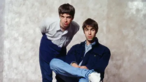 Koh Hasebe/Shinko Music/Getty Images Noel Gallagher standing, hunch-backed, next to Liam Gallagher, cross-legged, each unsmiling as they stare at the camera.
