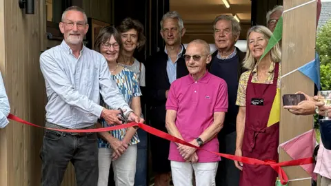 Villagers gather in a doorway to cut a ribbon to mark the official opening of the rebuilt village shop. There is a long red ribbon being cut by one man while other people are looking on. 