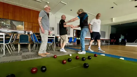 MARTIN GILES/BBC A group of four men playing short mat bowls at the Montagu working man's club in Huntingdon. Shane is on the right wearing a white t-shirt and black shorts. Two men are shaking hands and congratulating themselves in the middle. A man on the right is looking at the bowls on the floor, as is Shane. There are velvet covered function chairs in the background, and the bar shutters are down. There is a small stage to the right.