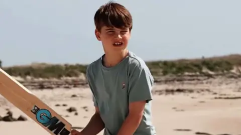 Prince Louis squints while holding a cricket bat on a beach