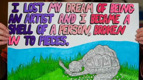 Peterborough City Council A poster depicts a person with a tortoise-like shell on their back while the are crouched in the grass. They lack on colour. Words on the poster read: "I lost my dream of being an artist and I became a shell of a person, broken into pieces."