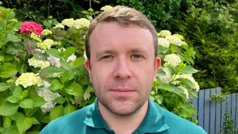 Peter McReynolds wearing a green polo shirt. Behind him are shrubs and flowers, and a blue wooden fence. 