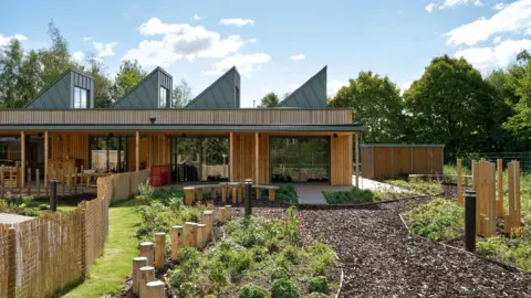 Daniel Hopkinson A wooden building that has large glass windows and a garden with a bark path and plants all around.