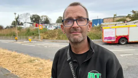 Jonathan Harvey outside Locking Farm Industrial Estate in Weston-super-Mare. He has short dark hair and stubble. He is wearing glasses with thick black frames and a black jumper with a green logo on the chest.