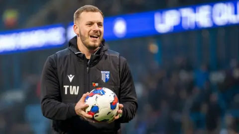 Gillingham FC A man with light brown hair and wearing a black jacket, is holding a football. He is standing inside a stadium.