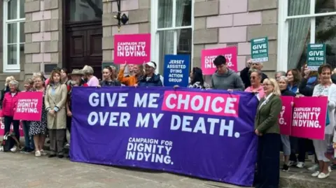 Campaigners for assisted dying with banners and placards in Jersey standing outside a building.