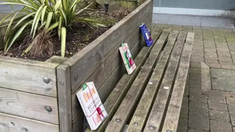 Three books, on a wooden bench. The books are spaced out evenly and leaning again a plant box that is made of wood that the bench is attached to. The books are tied in red string and have green bookmarks that say 'free book' on them. There is a palm plant in the plant box. 