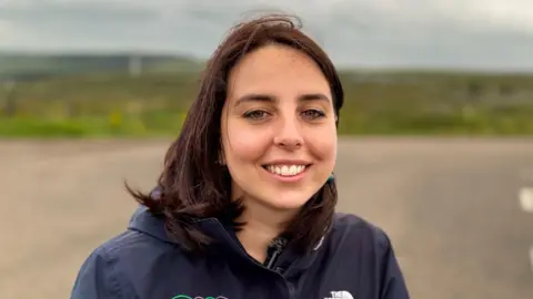 A woman with long brown hair, smiling at the camera. She is wearing a navy rain jacket.