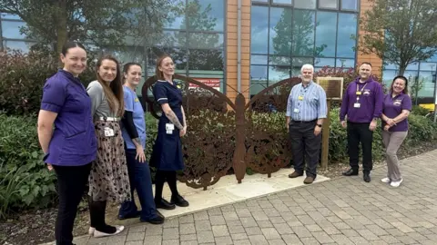 Northwest Anglia Foundation NHS Trust Seven people including five women and two men are standing on either side of a brown butterfly sculpture. 