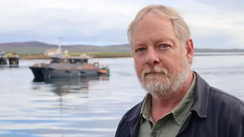 Peter Ferguson-Smyth, a light-haired man with a beard, looks at the camera from a harbour area. There is a body of water behind him and a small boat Passes by behind him.