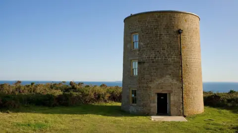 A stone tower with two windows in a field. In the background is the sea on a clear sky.