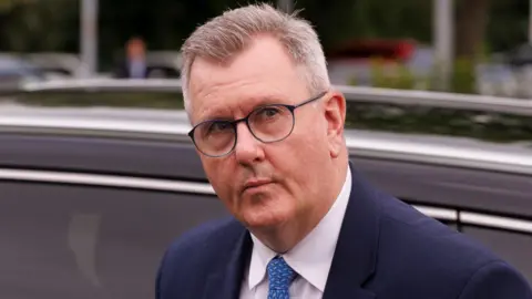 A man with short, mostly grey hair, is exiting a car. He is wearing black-rimmed glasses, a blue suit jacket, white shirt and blue-patterned tie. The car behind him is black. Out of focus, in the background, are other vehicles, lampposts and shrubbery. 