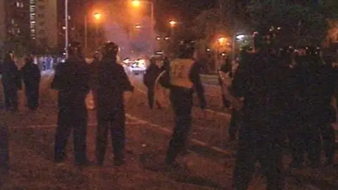 A grainy photo from 1995 showing a large amount police officers wearing armour and stood together looking at a car on fire