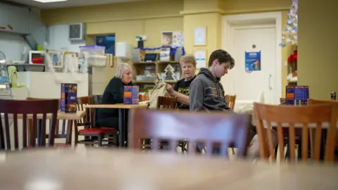 Interior view of Andy's Coffee House shows two women sitting at one table and a young man at another. The walls are light yellow and there are tinsel decorations pinned around them. We see menus on the wooden tables and a counter at the back of the room.