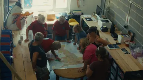 Braving the Blue - Our Lives, BBC Cymru Wales The six women standing around a table looking at a large map of the United Kingdom. They are joined by Sian's daughter who works for the RNLI and a woman from their support team. Sian has a ruler and is measuring the distance between Ireland and Wales. They are standing in the Blue Tit Chill Swimmers head quarters.