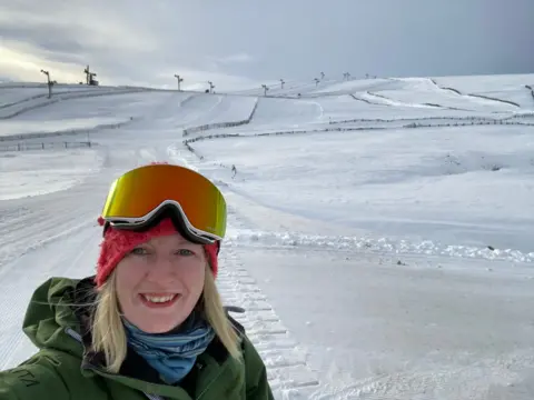 Annette Cockburn Annette Cockburn, who has ski goggles perched on top of her head, stands at the bottom of snow-covered ski slope. She is wrapped up in a blue scarf and a green jacket. She has blonde hair and is smiling at the camera in this selfie