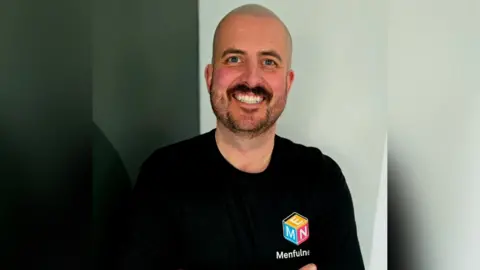 Jane Bowes/BBC Jack Woodhams smiles at the camera. he has a shaved head and a short, dark beard and wears a black t-shirt with the Menfulness logo on the chest - a cube with blue, orange and pink sides and white lettering below spelling out Menfulness.