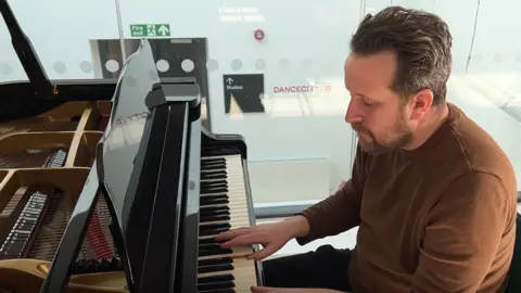 Barry Hyde sitting at a grand piano, with his fingers on the keys. He is wearing a brown top with the sleeves rolled up