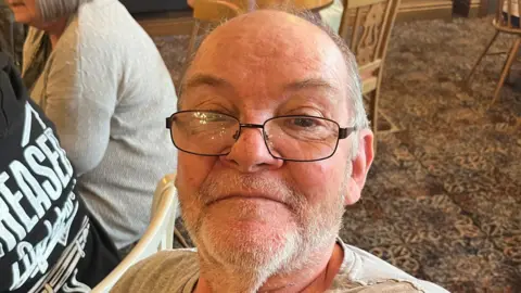 John Devine/BBC A man looking straight a the camera with a neutral expression. He is wearing glasses. There are two people sitting on wooden chairs behind him.