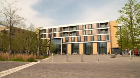 Cambridge City Council A modern beige building with big windows and wooden paneling. In front is a pedestrianised area with chrome bollards and trees surrounding it.