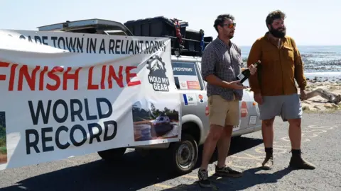 Two men stand in front of a parked three-wheel car. One of the man is holding a bottle of champagne. A very large white sign is on display behind them and reads London to Cape Town in a Reliant Robin. Finish Line World Record. 