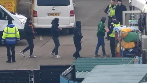 Getty Images A group of people thought to be migrants are brought in to Dover, Kent, by the RNLI Dungeness Lifeboat following a small boat incident in the Channel. They are pictured crossing the road at the Port of Dover.